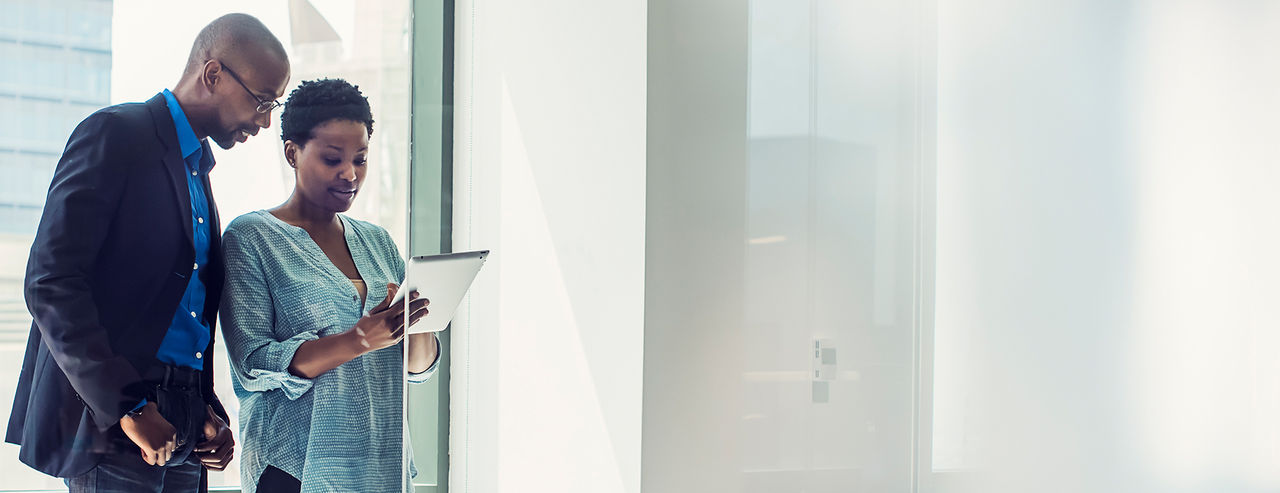 A man and woman standing by a city building window while looking at a tablet